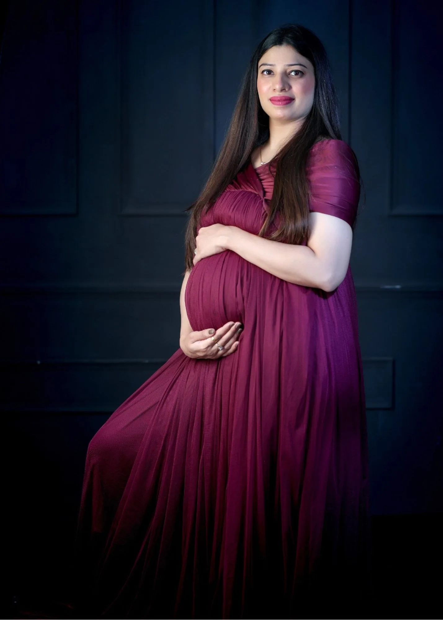 Expecting mother sitting in a rich maroon gown holding her baby bump