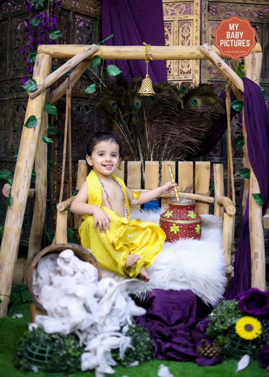 Little girl in a yellow Krishna dress smiling on a decorated jhula (swing), Janmashtami Krishna theme baby photoshoot in Delhi NCR.