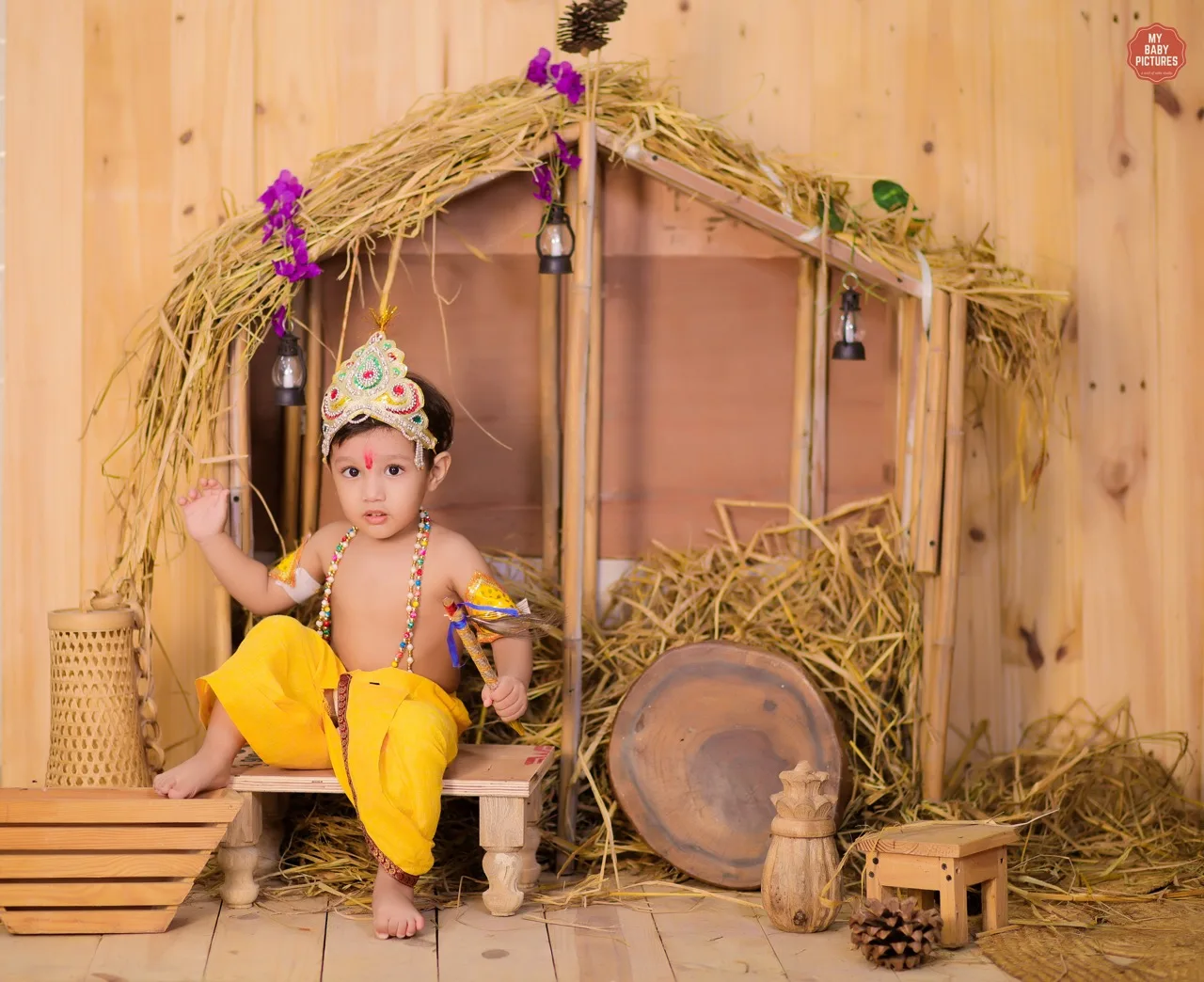 A waving baby boy dressed as Krishna in a straw hut studio setup, Janmashtami Krishna theme baby photoshoot in Delhi NCR.